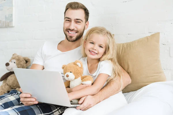 Glücklicher Vater und süße kleine Tochter benutzen Laptop zusammen und lächeln in die Kamera im Schlafzimmer — Stockfoto