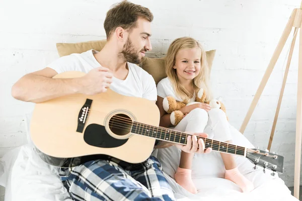 Feliz padre con guitarra acústica mirando linda hija pequeña con oso de peluche en la cama - foto de stock