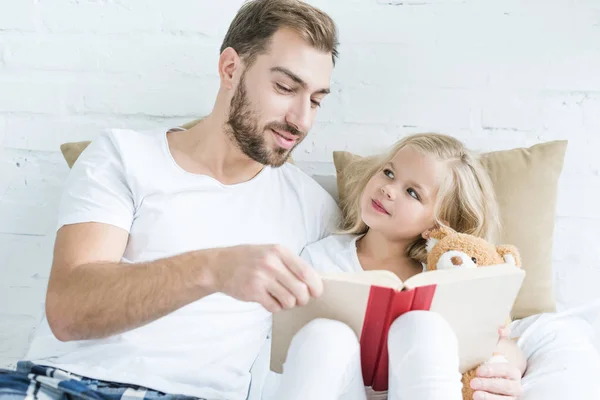 Father and cute little daughter with teddy bear reading book together on bed — Stock Photo