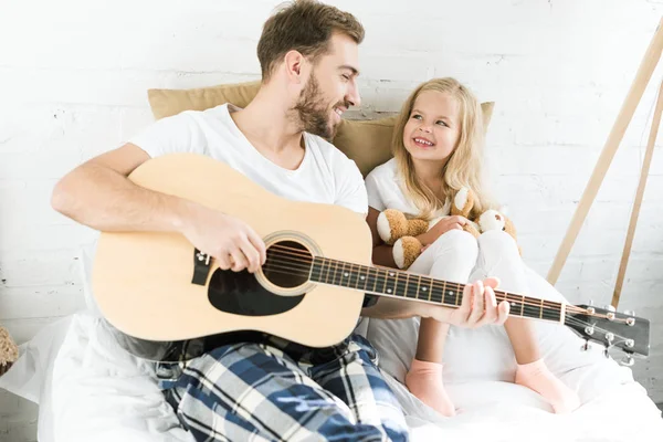 Feliz padre con guitarra acústica y linda hija pequeña con oso de peluche sonriendo el uno al otro en casa - foto de stock