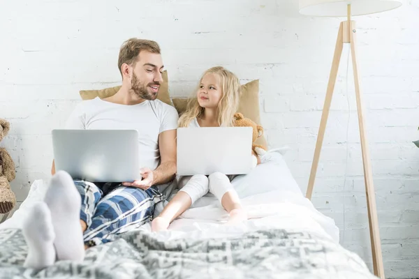 Glücklicher Vater und Tochter lächeln einander an, während sie Laptops auf dem Bett benutzen — Stockfoto