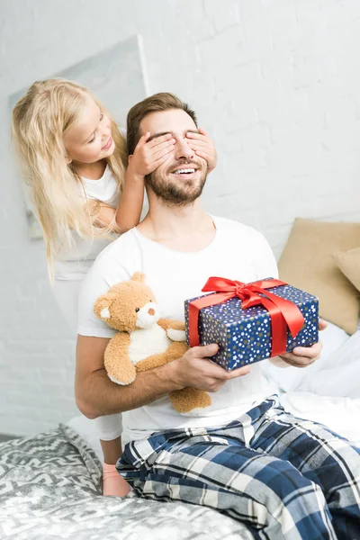 Entzückende kleine Tochter schließt Augen vor glücklichem Vater mit Geschenkbox und Teddybär — Stockfoto