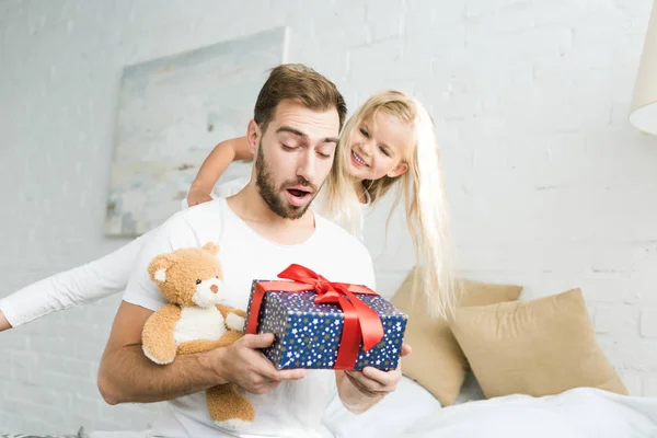 Adorable pequeña hija feliz mirando sorprendido padre sosteniendo caja de regalo y osito de peluche - foto de stock