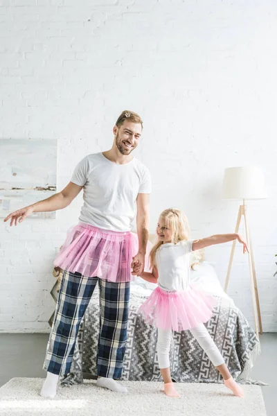 Счастливый отец и очаровательная маленькая дочь в розовых юбочках в пачках, держась за руки и танцуя дома — стоковое фото