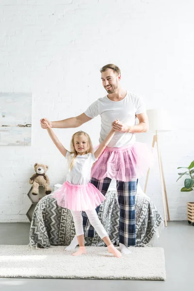 Щасливий батько і дочка в рожевих спідницях, тримаючись за руки і танцюючи разом вдома — стокове фото