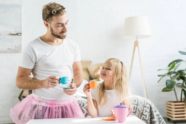Счастливый отец в розовой юбке в пачке смотрит на милую маленькую дочь, притворяющуюся, что у нее дома чаепитие — стоковое фото