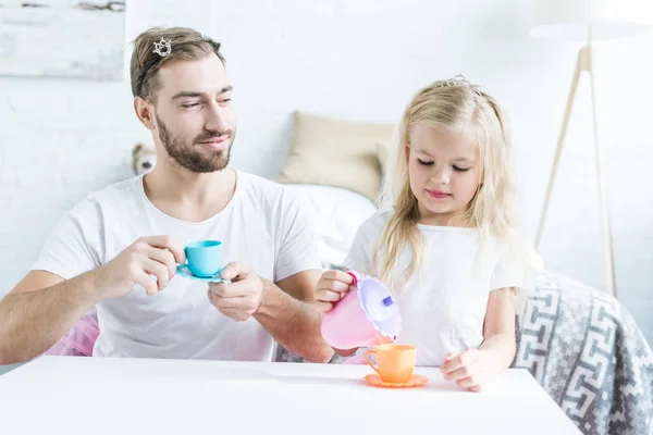 Souriant père tenant jouet tasse et regardant petite fille verser le thé de bouilloire jouet — Photo de stock