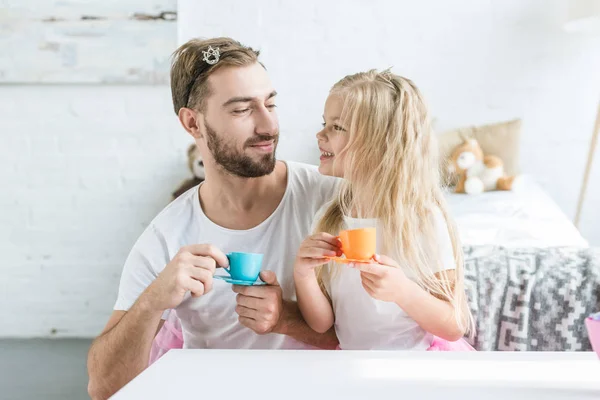 Счастливые отец и дочь улыбаются друг другу, играя вместе и притворяясь, что чаепитие — стоковое фото
