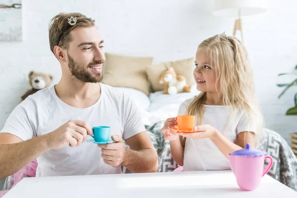 Feliz padre e hija sonriendo el uno al otro mientras juegan al té en casa - foto de stock