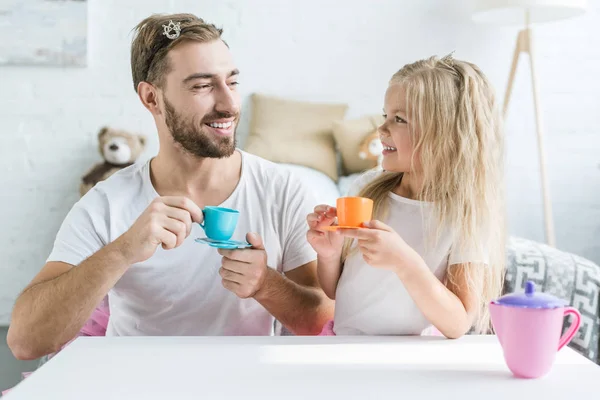 Feliz padre e hija sonriéndose mientras juegan con platos de juguete en casa - foto de stock