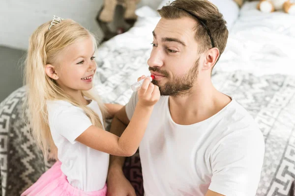 Adorable little daughter applying red lipstick to happy bearded father at home — Stock Photo