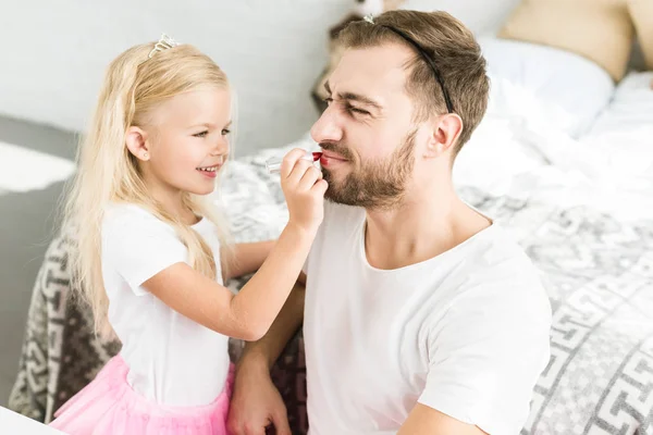 Niedliche kleine Tochter bringt zu Hause glücklichen bärtigen Vater mit rotem Lippenstift an — Stockfoto