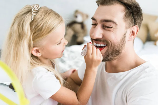 Cute little daughter applying red lipstick to happy bearded father — Stock Photo