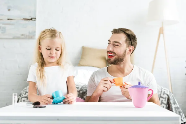 Pai feliz com maquiagem e filhinha fofa sorrindo um ao outro enquanto brincava com pratos de brinquedo em casa — Fotografia de Stock