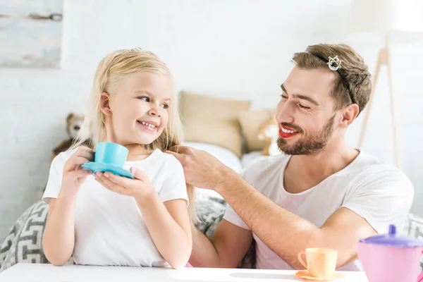 Feliz padre trenza trenzado a adorable hija fingiendo tener fiesta de té en casa - foto de stock