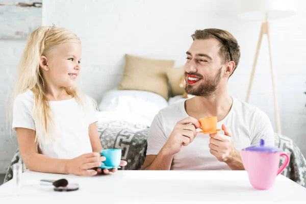 Padre con lápiz labial rojo y linda hija feliz sonriéndose mientras juegan con platos de juguete en casa - foto de stock