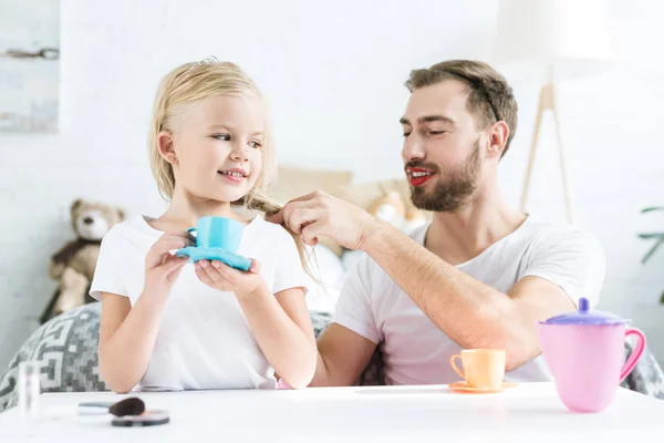 Glücklicher Vater flechtet Zopf zu lächelnder Tochter und gibt vor, zu Hause Tee zu trinken — Stockfoto