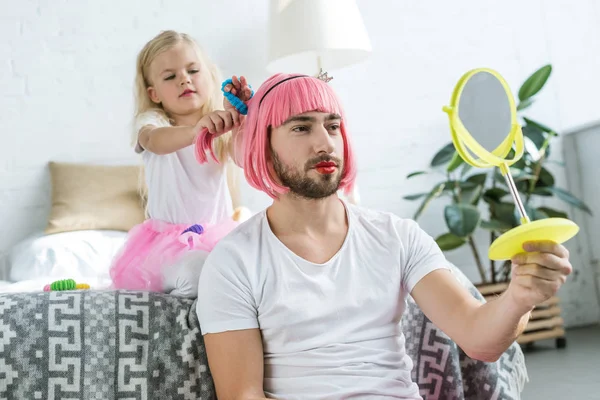 Чарівна маленька дочка в спідниці tutu грає з батьком в рожевій перуці, дивлячись на дзеркало — стокове фото