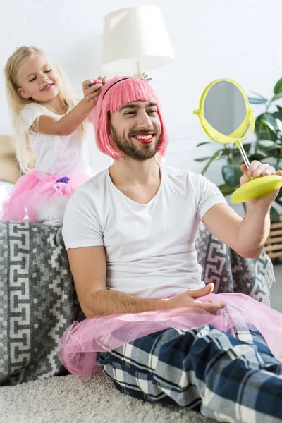 Niedliche kleine Tochter im Tutu-Rock spielt mit lächelndem Vater in rosa Perücke und schaut in den Spiegel — Stockfoto