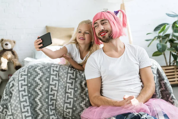 Heureuse petite fille et souriant père en perruque rose prendre selfie avec smartphone à la maison — Photo de stock