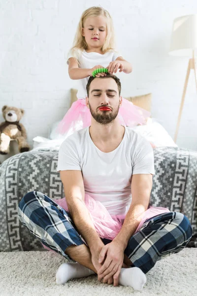 Adorable little daughter in pink tutu skirt doing hairstyle to father with red lipstick — Stock Photo