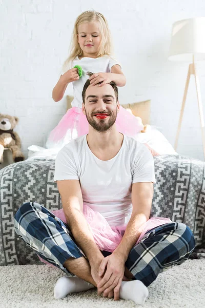 Adorable little daughter in pink tutu skirt doing hairstyle to happy father with red lipstick — Stock Photo
