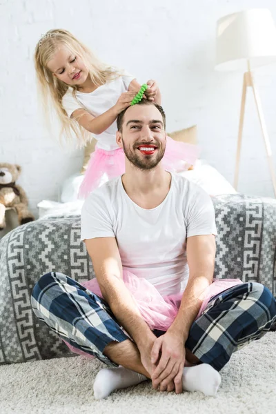 Cute little daughter in pink tutu skirt doing hairstyle to happy father with red lipstick — Stock Photo