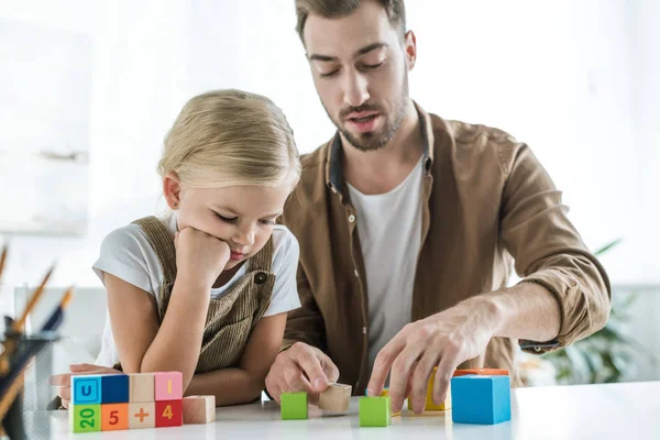 Pai e pequena filha entediada aprendendo matemática com cubos coloridos em casa — Fotografia de Stock