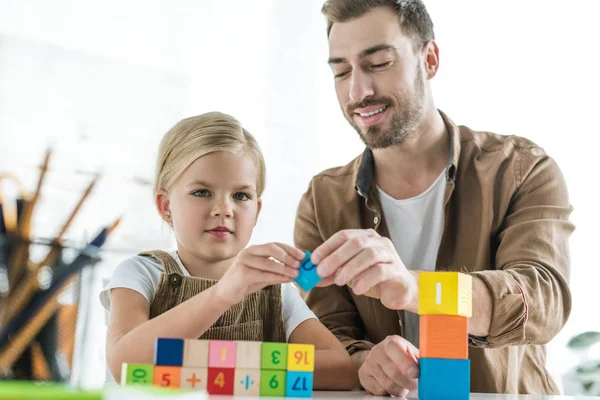 Padre sonriente y linda hija pequeña aprendiendo matemáticas con cubos de colores en casa - foto de stock