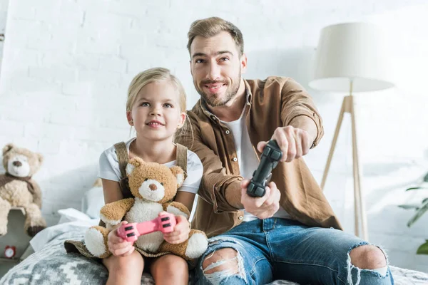 Feliz padre y linda hijita con oso de peluche jugando con joysticks en casa - foto de stock