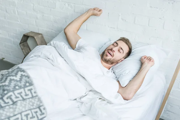 Alto ángulo vista joven hombre bostezando y estirándose en la cama - foto de stock