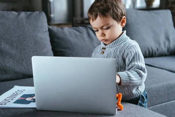 Messa a fuoco selettiva del bambino in piedi vicino al computer portatile e al giornale commerciale sul divano a casa — Foto stock