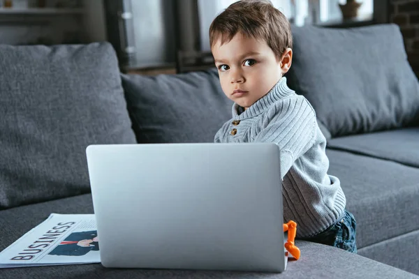 Kid standing near laptop and business newspaper on sofa at home — Stock Photo