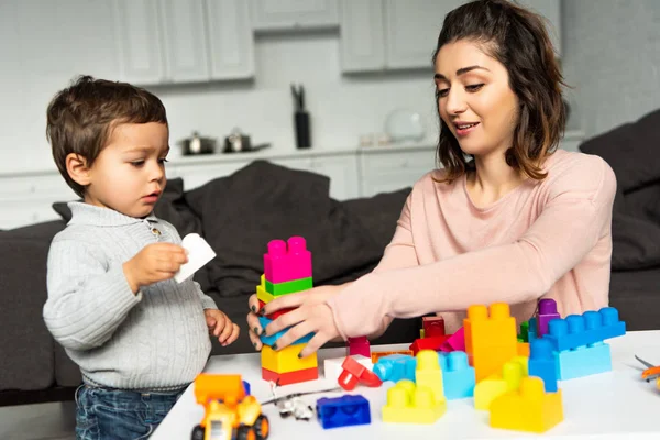 Atractiva mujer feliz y su pequeño hijo jugando con bloques de plástico de colores en casa - foto de stock