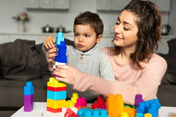 Улыбающаяся мать и маленький сын играют с красочными пластиковыми блоками дома — стоковое фото