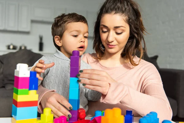 Menino bonito e sua mãe brincando com blocos de plástico coloridos em casa — Fotografia de Stock