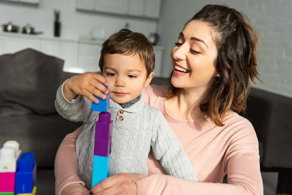 Menino alegre e sua mãe brincando com blocos de plástico coloridos em casa — Fotografia de Stock