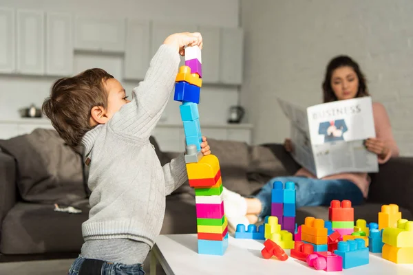 Foco seletivo de menino brincando com blocos de plástico coloridos, enquanto sua mãe está sentada atrás em casa — Fotografia de Stock