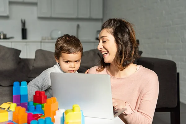 Niño y su madre que ríe usando el ordenador portátil en la mesa en la sala de estar en casa - foto de stock