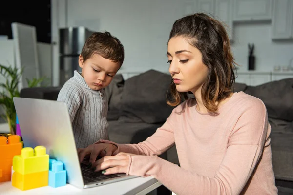 Foco seletivo do menino olhando para sua mãe usando laptop à mesa na sala de estar em casa — Fotografia de Stock