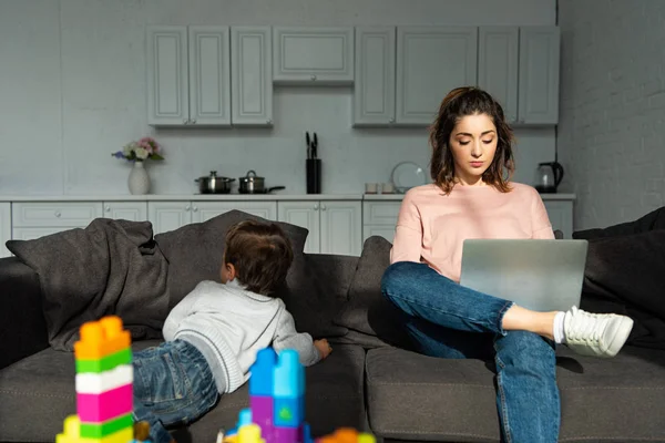 Visão traseira do menino subindo no sofá, enquanto sua mãe usando laptop em casa — Fotografia de Stock