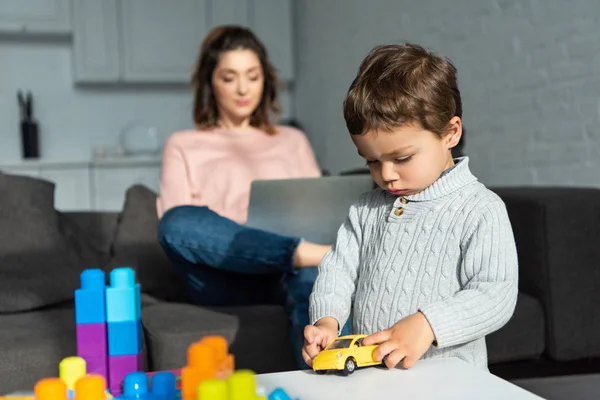 Child playing with toy car while his mother using laptop behind at home — Stock Photo