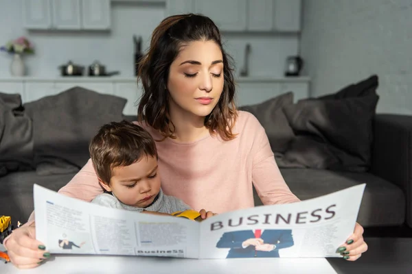 Hermosa mujer y su pequeño hijo leyendo periódico de negocios en la sala de estar en casa - foto de stock