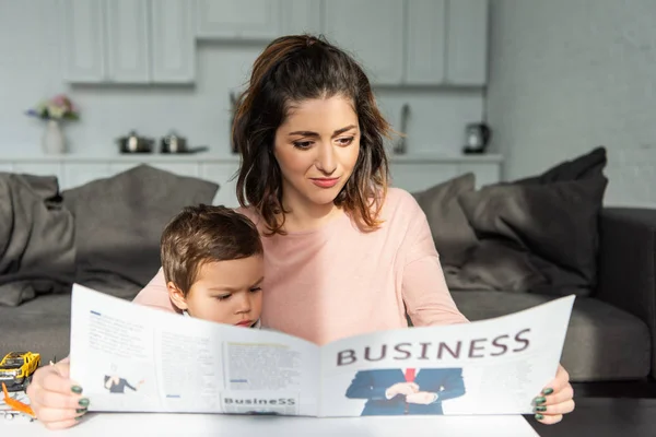 Красивая женщина и ее маленький сын читают деловую газету в гостиной дома — стоковое фото