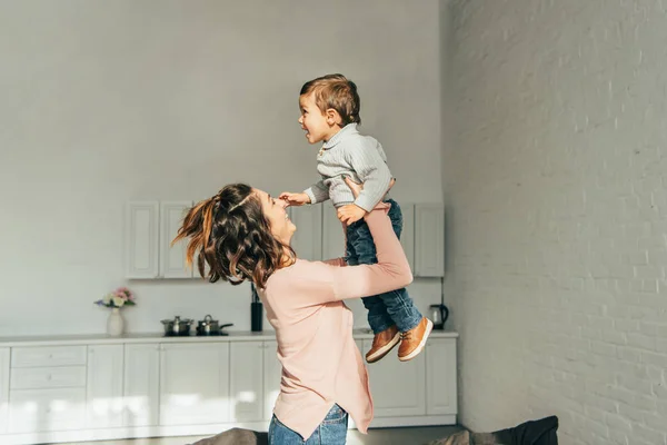 Seitenansicht einer lächelnden Frau, die ihren entzückenden Sohn zu Hause im Wohnzimmer aufzieht — Stockfoto