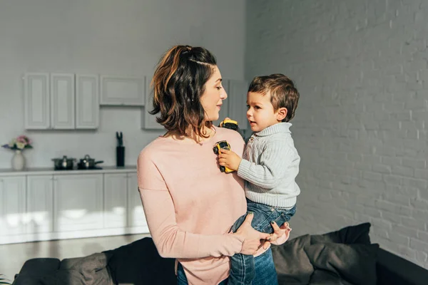 Attraktive Mutter hält kleinen Sohn mit Spielzeugautos auf Händen zu Hause — Stockfoto