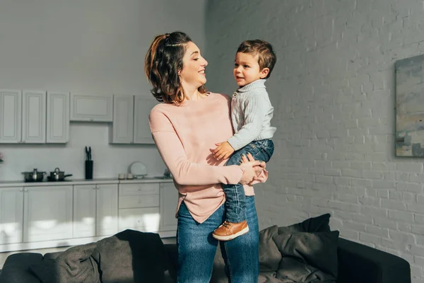 Attraktive, glückliche Frau, die ihren entzückenden kleinen Sohn zu Hause auf Händen hält — Stockfoto