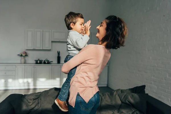 Vista lateral de la madre sonriente sosteniendo adorable pequeño hijo en las manos en casa - foto de stock