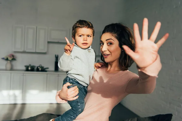 Feliz atractiva mujer sosteniendo pequeño hijo y saludando a mano en casa - foto de stock
