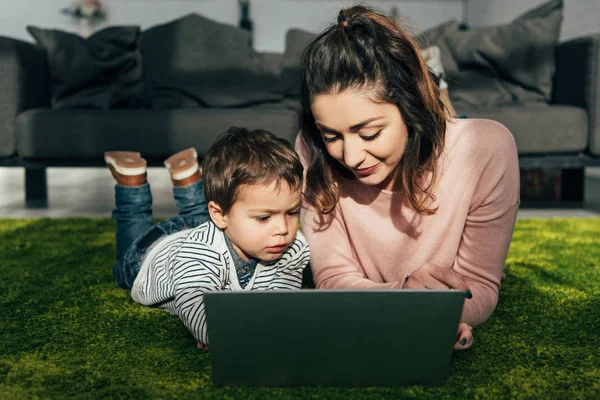 Mãe sorridente e pequeno filho deitado no chão com laptop em casa — Fotografia de Stock